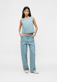 Femme portant un haut sans manches en denim bleu clair et un jean taille haute à jambes larges, les mains dans les poches, debout sur un fond blanc.