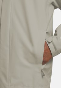 Lichtbeige jas met een gladde textuur, voorzien van een ritszak en verstelbare manchetten. Minimalistisch ontwerp, versterkte stiksel details.