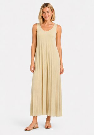 Vrouw draagt een mouwloze beige geplooide maxi-jurk en platte sandalen, staand met een lichte glimlach op een effen achtergrond.