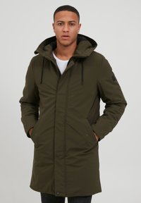 Abrigo parka verde oliva con capucha, bolsillos frontales y cierre de cremallera. La tela es suave y ligeramente texturizada, manteniendo una silueta ajustada.