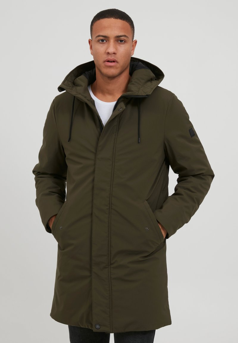 Abrigo parka verde oliva con capucha, bolsillos frontales y cierre de cremallera. La tela es suave y ligeramente texturizada, manteniendo una silueta ajustada.
