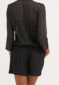Top negro transparente con mangas tres cuartos, que presenta patrones verticales de hilo plateado. Combinado con pantalones cortos negros, de textura suave y ajuste casual.