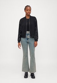 Veste bomber noire avec des manches couvertes de sequins, fermeture éclair à l'avant et poignets côtelés. Portée sur un haut noir et associée à un jean évasé bleu clair.