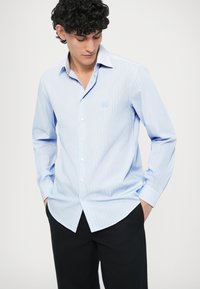 Světle modrá košile na knoflíky s vertikálními pruhy, dlouhými rukávy a zaobleným lemem. Na hrudi má malý vyšívaný logo.