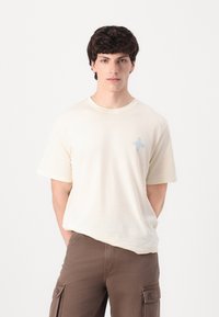 Camiseta de color beige claro con cuello redondo y mangas cortas. Presenta un pequeño diseño de flor azul en la parte izquierda del pecho. Textura de algodón suave.