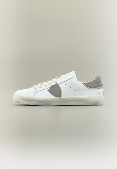 Baskets basses blanches avec lacets crème, languette de talon grise, patch latéral gris, et semelle en caoutchouc blanche légèrement usée sur un fond neutre.