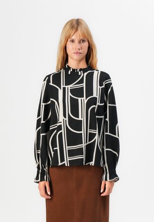 Woman wearing a black long-sleeve blouse with white geometric lines and a brown skirt, standing against a plain white background.