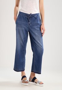 Pantalon large en denim bleu avec une taille élastique et un cordon de serrage. Comprend des poches latérales et un ourlet effiloché. Assorti à des sandales à plateforme.