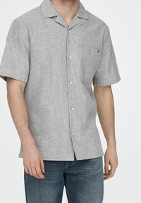 Man med kortärmad, knappad randig skjorta med bröstficka, tillsammans med blå jeans, mot en enkel bakgrund.