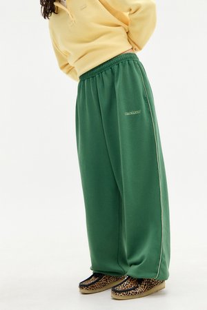 Grüne weite Sweatpants mit seitlichen Paspeln, ausgestattet mit einem elastischen Bund und einem kleinen gestickten Logo. Kombiniert mit braunen Schuhen im Leopardenmuster.