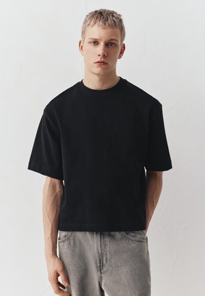 Jeune homme aux cheveux blonds courts portant un T-shirt noir ample et un jean gris clair, debout devant un mur blanc texturé.
