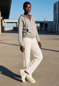 Mujer con sudadera con cremallera gris, pantalones blancos y zapatillas blancas, de pie al aire libre en una plaza de hormigón soleada con edificios modernos al fondo.