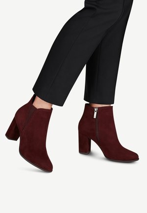 Femme portant des bottines en daim rouge foncé avec talons carrés et fermetures éclair latérales, associées à un pantalon noir court.