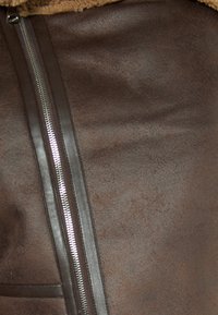 Brązowy materiał z faux skóry o gładkiej fakturze, z wyraźnym srebrnym zamkiem błyskawicznym po boku, uzupełniony akcentem w kolorze wełnianym jasnobrązowym.