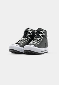 Schwarze High-Top-Sneaker aus synthetischem Material mit weißer Gummisohle und Schnürsenkeln, ausgestattet mit einer Schlaufe an der Ferse und einem strukturierten Muster auf der Sohle.