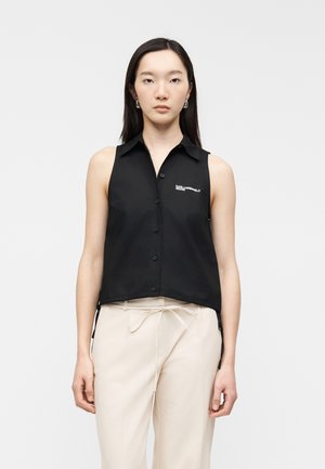 Noor naine, kes kannab musta varrukateta nööbitavat särki rinnal logo ja heledaid beežipükse, seisab ühevärvilise tausta ees.