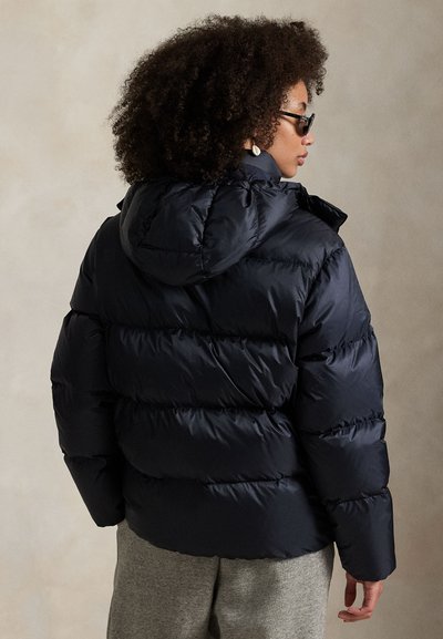 Vrouw met krullend haar, zonnebril en zwarte pufferjas met capuchon, staand tegen een effen beige achtergrond.