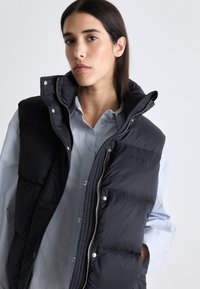 Svart puffer vest med høy krage, med frontglidelås og trykknapper, båret over en lyseblå skjorte. Glatt tekstur, rutet design.