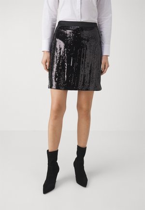 Femme portant une mini-jupe noire scintillante à sequins associée à une chemise blanche à manches longues et des bottines noires.