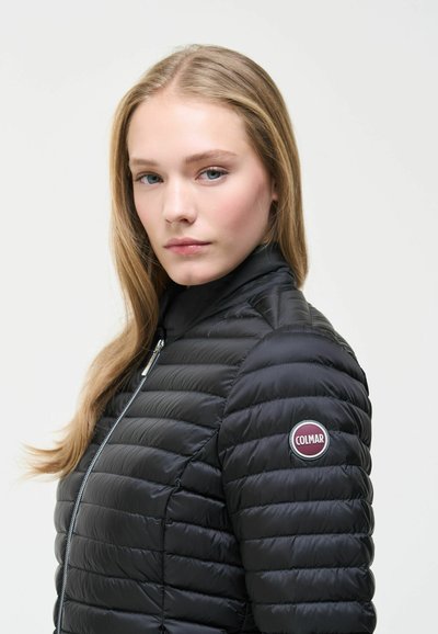 Jeune femme aux longs cheveux blonds portant une veste matelassée noire Colmar avec une fermeture éclair et un logo sur la manche gauche.