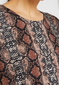 Femme portant un haut à col rond en tissu avec un motif de peau de serpent brun, noir et beige.