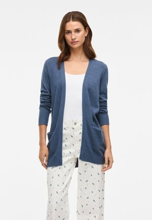Cardigan de manga longa, aberto à frente, em tecido de malha azul com punhos e bainha canelados; possui dois bolsos frontais. Usado sobre uma blusa branca e calças com padrão.