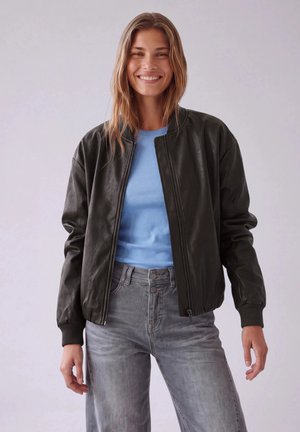 Une femme souriante porte une veste en cuir noire par-dessus une chemise bleue et un jean gris, sur un fond uni.