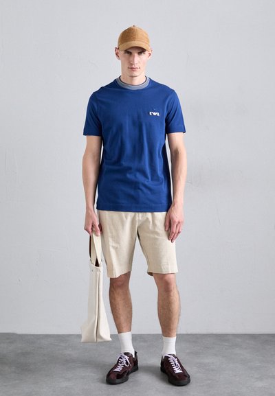 T-shirt bleu à manches courtes avec logo, shorts beiges, chaussettes blanches et baskets noires. Le mannequin tient un sac fourre-tout beige, debout sur une surface grise lisse.