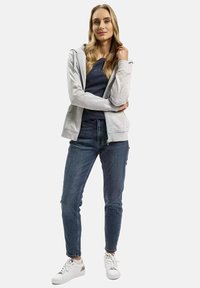 Szary rozpinany hoodie na ciemnoniebieskiej bluzce, w połączeniu z ciemnymi dżinsami i białymi sneakersami. Model stoi w luźnej pozie, z rękoma skrzyżowanymi. Gładki materiał.