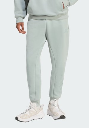 Personne portant un pantalon de jogging vert clair et des baskets blanches, debout avec une main dans la poche sur un fond uni.