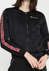 Svart bomberjacka med dragkedja framtill, ribbade ärmslut och logotyp på bröstet. Sidosträck har växlande röda och vita designdetaljer.