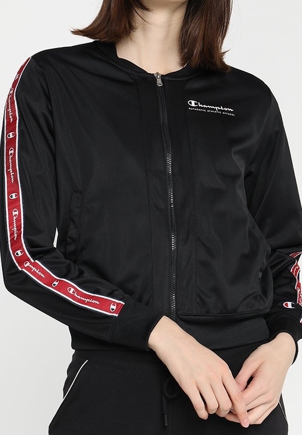 Svart bomberjacka med dragkedja framtill, ribbade ärmslut och logotyp på bröstet. Sidosträck har växlande röda och vita designdetaljer.