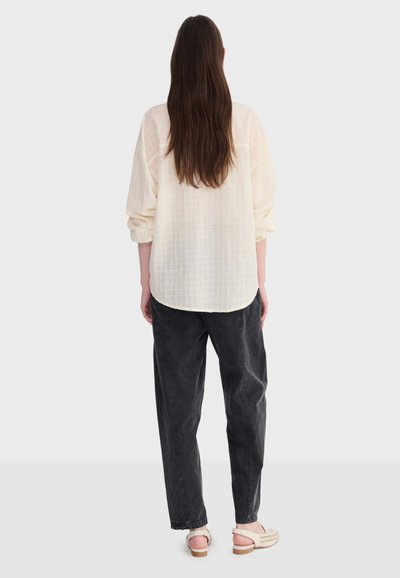 Femme debout, de dos, portant une chemise texturée crème à manches longues, un pantalon noir ample et des sandales beige sur un fond uni.