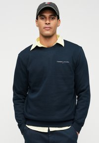 Sweatshirt azul-marinho com decote arredondado e punhos canelados, apresentando um logótipo branco "TOMMY JEANS". Usado por cima de uma camisa amarela com gola.