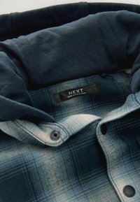 Närbild av kragen på en blårutig skjorta med mjukt navy-färgat innerfoder och en svart etikett som står "NEXT Made in Bangladesh."