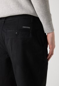 Černé kalhoty s zadní kapsou s knoflíkovým zapínáním a štítkem Calvin Klein. Materiál vypadá hladce a má strukturovaný střih.