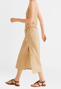 Kvinde går iført en beige og hvid lodret stribet ærmeløs kjole med slidser i siden og sorte stroppede flade sandaler.