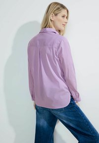 Long-sleeve shirt in light purple with white vertical stripes, featuring a pointed collar and a back pleat, paired with blue denim jeans.