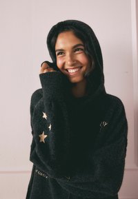 Schwarzer Kapuzenpullover aus weichem Material, mit goldenen Sternelementen an den Ärmeln und einem lässigen, oversized Design.