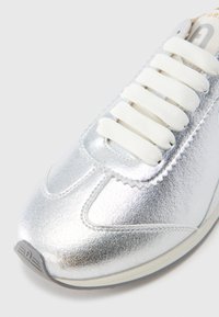 Ασημί μεταλλικό αθλητικό παπούτσι με ομαλή υφή, λευκά επίπεδα κορδόνια, στρογγυλή μύτη και διακριτική λεπτομέρεια ζιγκ-ζαγκ ραφής κατά μήκος των άκρων.