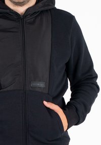 Felpa nera con cappuccio caratterizzata da una zip frontale, pannelli di tessuto testurizzato e una patch con logo sul petto. Include una tasca frontale e un cappuccio.