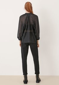 Blusa nera trasparente con motivo a pois. Caratterizzata da un collo arricciato e maniche lunghe. Abbinata a pantaloni neri attillati e stivaletti.