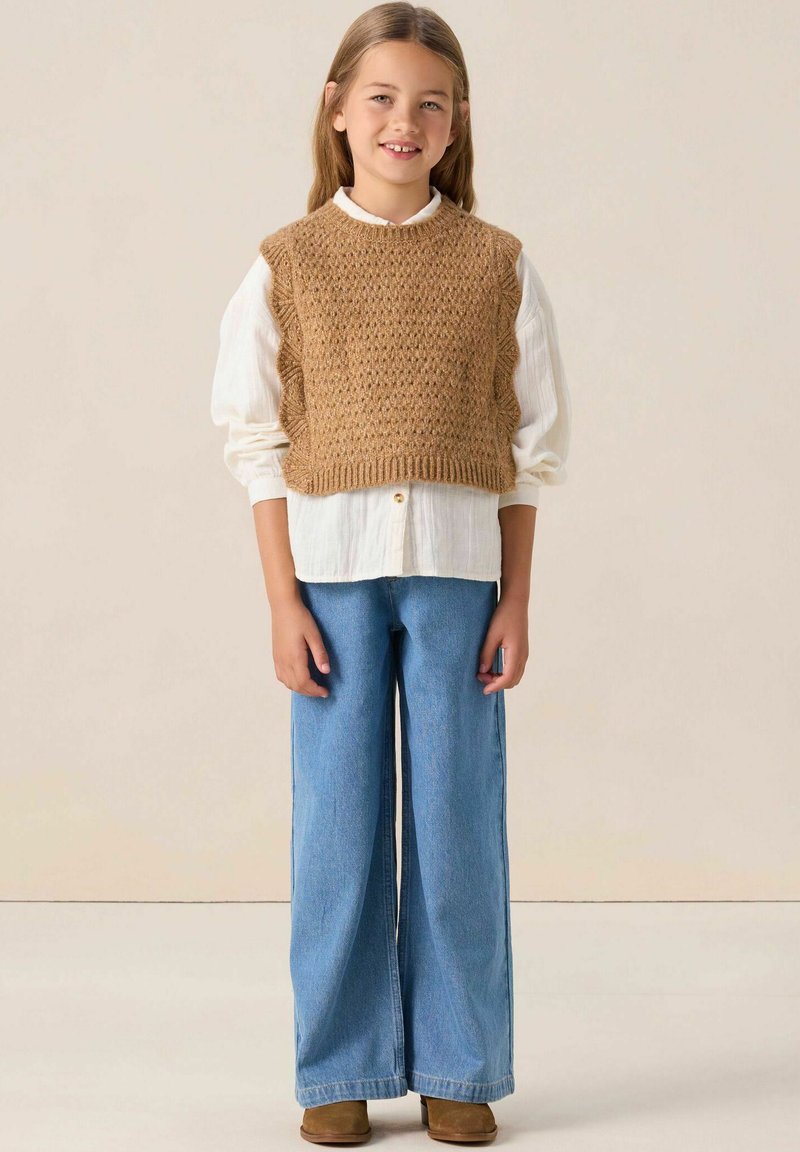 Un gilet en tricot marron par-dessus une blouse blanche, associé à un jean bleu décontracté et des bottes marron. Le gilet présente des détails texturés et un col rond.