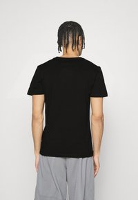 Man med flätat hår som bär en svart t-shirt och grå byxor, som står vänd mot en vit vägg.