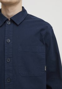 Navy blue button-up shirt made of cotton, featuring a collar, large front pocket, and five buttons along the placket.