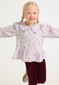 Blusa viola chiaro con stampa floreale, colletto a volant e chiusura con bottoni. Vita a peplo, maniche lunghe a sbuffo. Abbinata a pantaloni di velluto a coste color bordeaux.