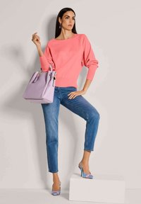 Roze pullover trui, blauwe spijkerbroek, lavendel handtas en blauwe hakken met een bloemdetail. Model poseert tegen een neutrale achtergrond.