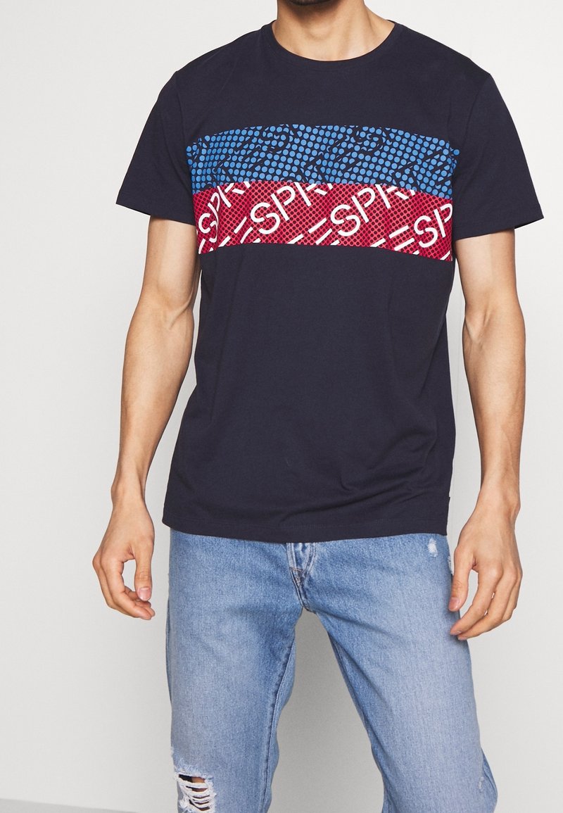 Homme portant un t-shirt noir avec des rayures horizontales à motifs bleus et rouges et un jean bleu clair déchiré, debout sur fond blanc.