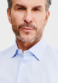 Jasnoniebieska koszula z guzikiem z wyraźnym kołnierzykiem, subtelną teksturą i widocznymi detalami szwów w pobliżu miejsca na guziki.