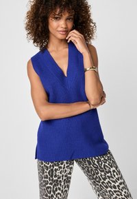 Blauw, mouwloos gebreid vest met een diepe V-hals en geribbelde textuur. Gecombineerd met grijs en zwarte panterprint broeken.